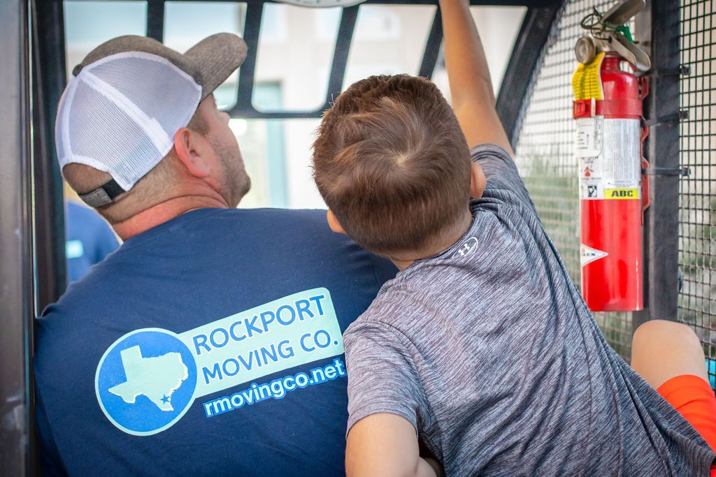 Man and child in a moving truck. Man wears a Rockport Moving Co. shirt. Child reaches up. Fire extinguisher visible.