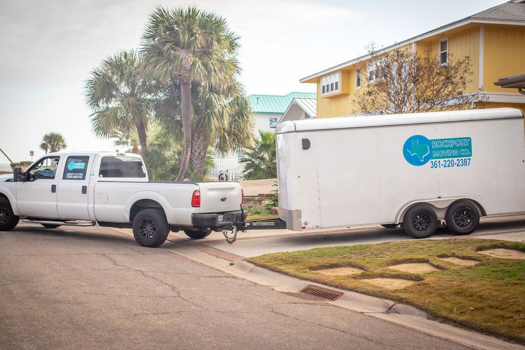 White truck towing a white trailer on a street next to a yellow house.