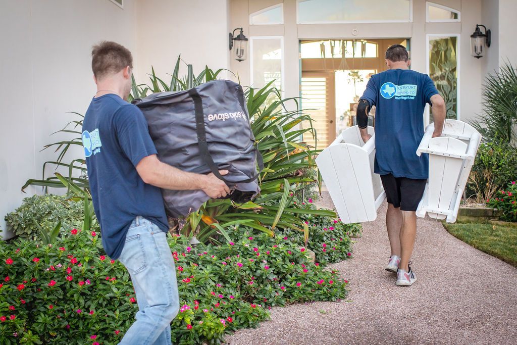 Two people carrying items out of a house, walking on a stone path. One holds a large bag, the other a white chair.