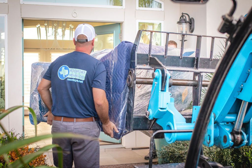 Man carrying wrapped furniture toward a house. Blue forklift, gray pants, blue shirt, white cap.