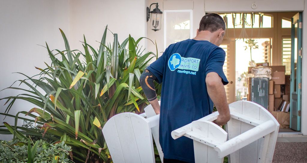 Man in blue shirt carrying white chairs outside a house with moving boxes.