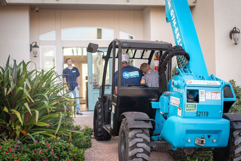 Blue forklift at a building entrance; workers are inside. One man watches. Lush green landscaping.