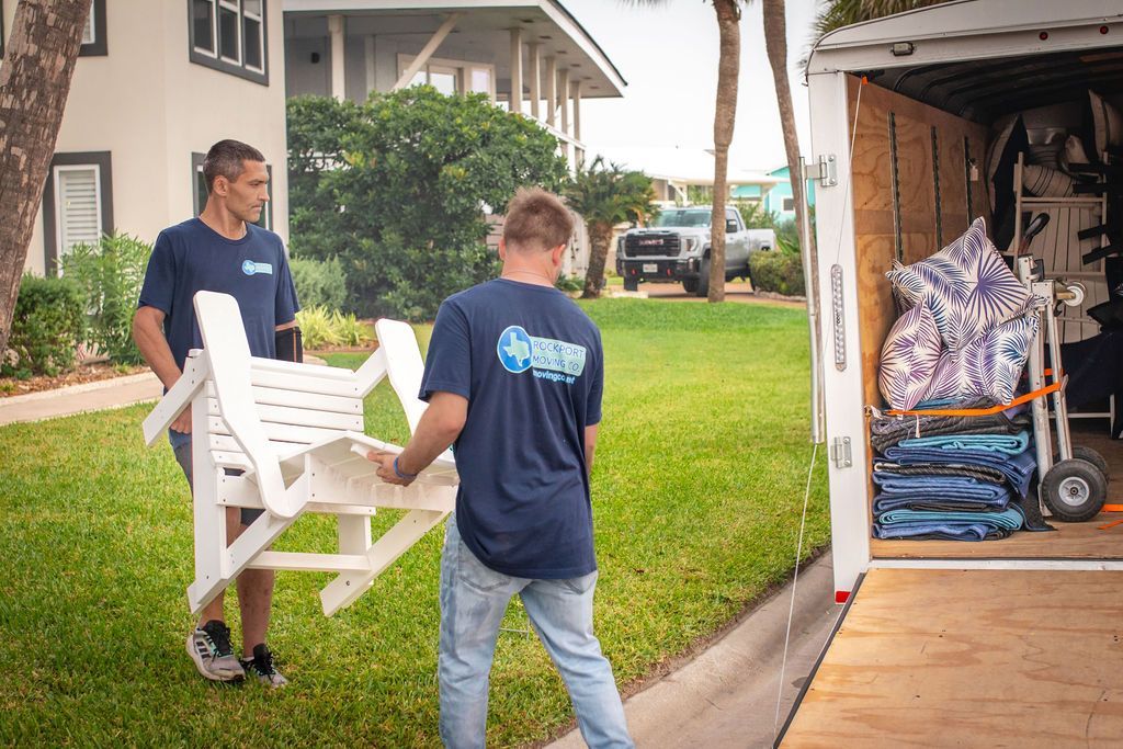 Two movers carrying a white chair towards a moving truck parked near a grassy lawn.