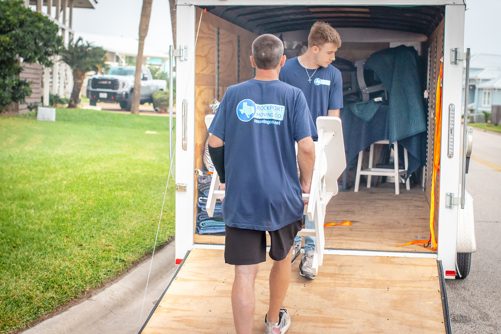 Two movers carrying furniture into a white trailer parked on a road with grass.