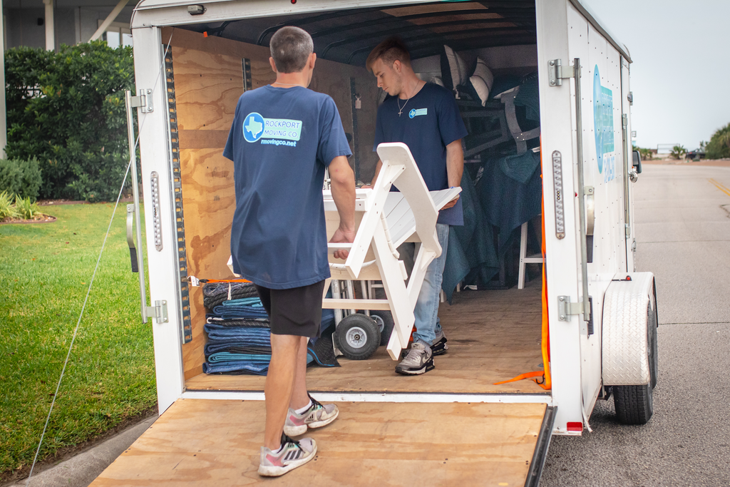 Two people loading a white folding chair into a trailer on a driveway.