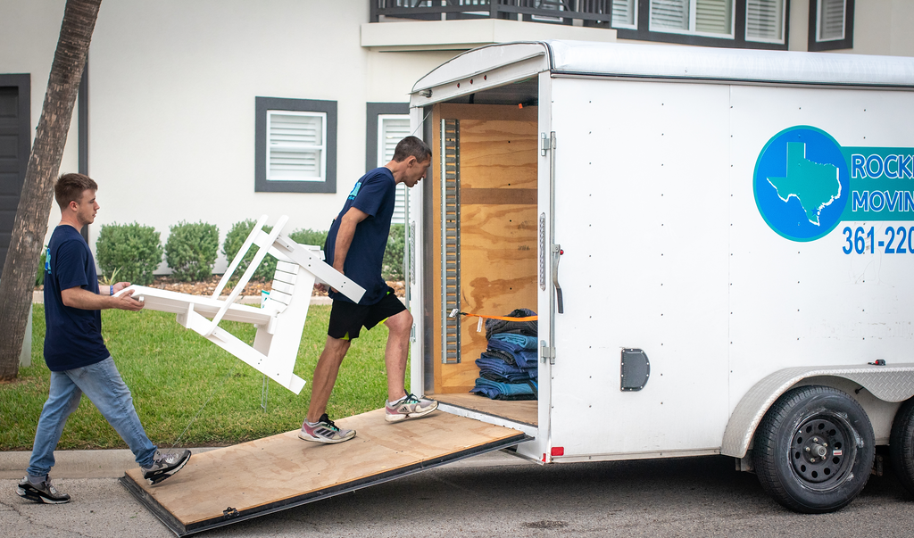 Two movers loading furniture into a white trailer with a ramp in front of a building.