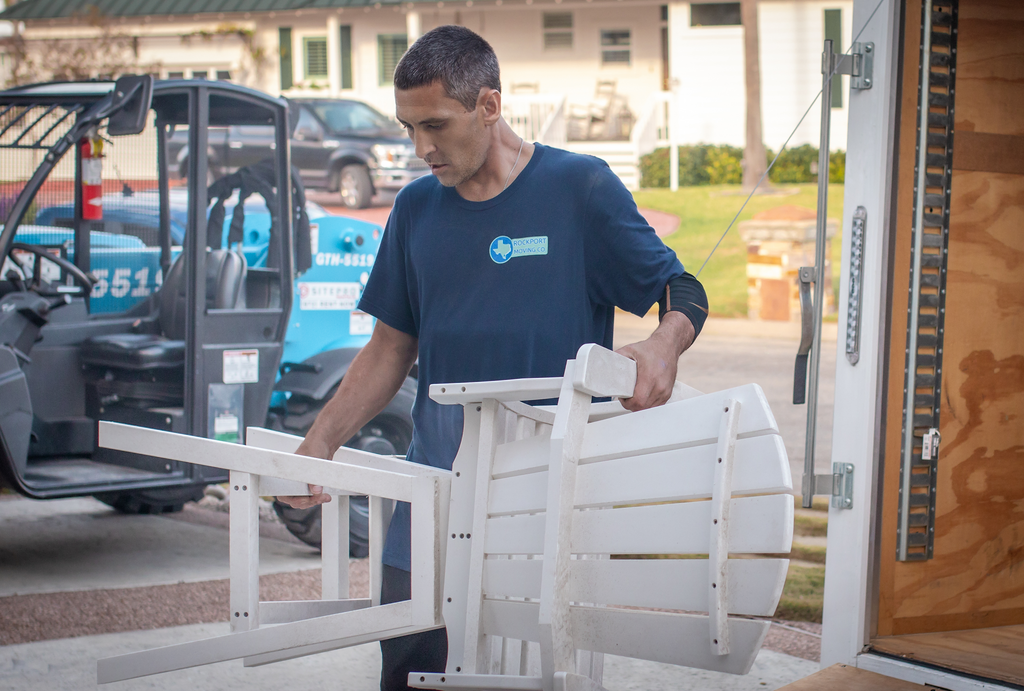 Man in blue shirt loading white chair into a truck. Outside, daytime.