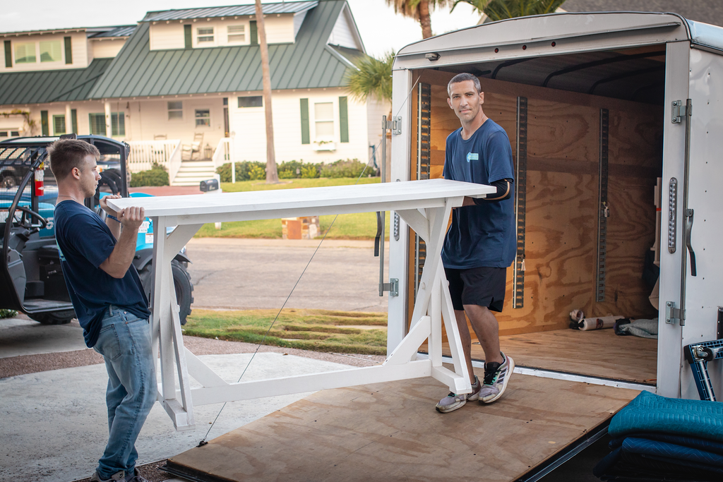 Two people loading a white table into a trailer; outside a house, on a ramp.