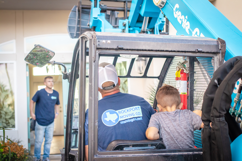 Man operating a Genie lift with a child. Another man watches near a building's entrance. Blue machinery.