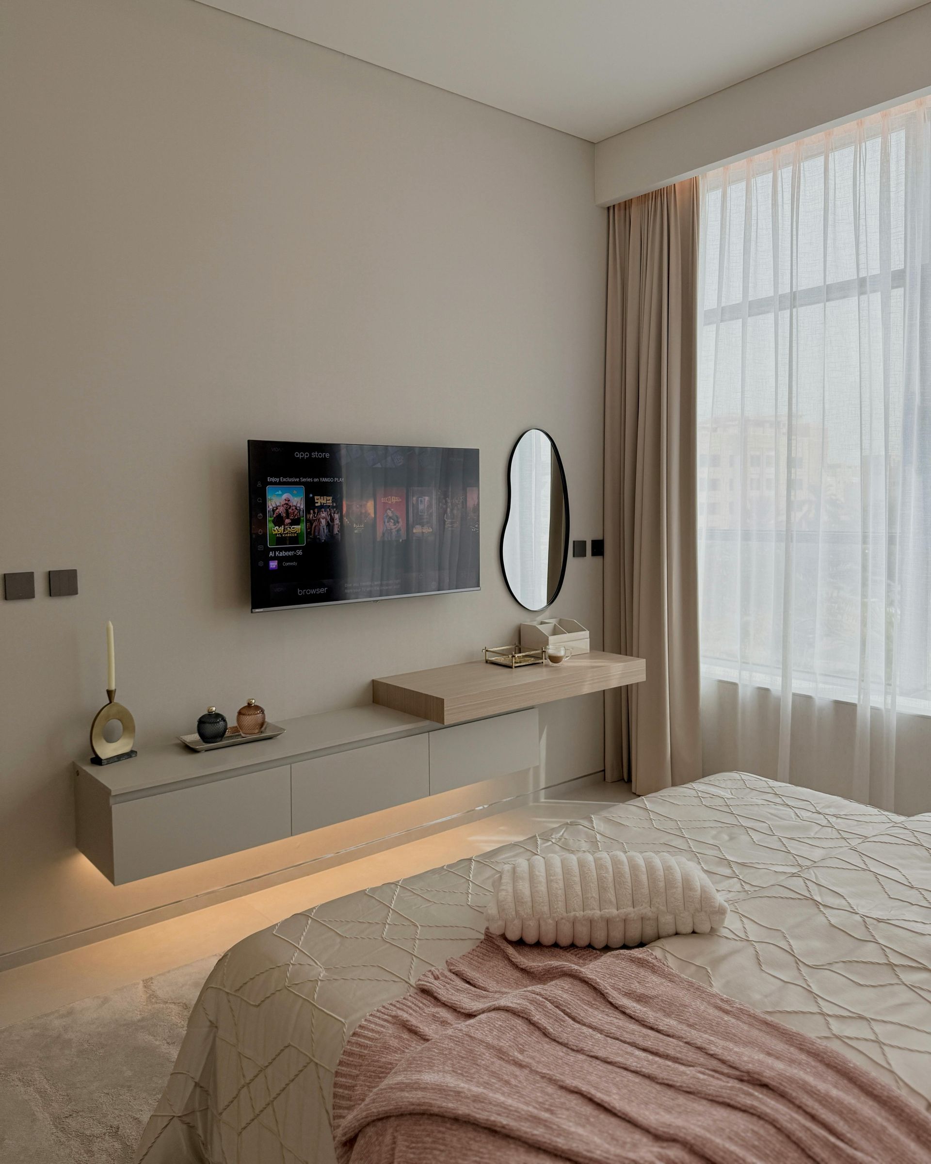 Bedroom with a floating TV and vanity, neutral colors, large window, bed with pink blanket.