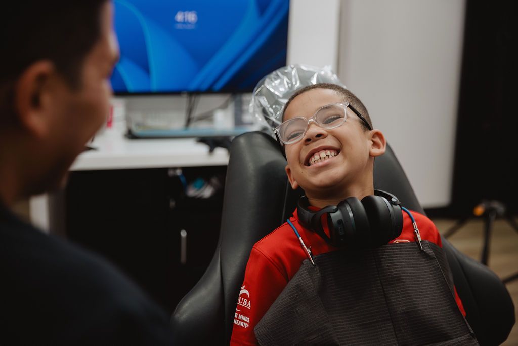 Boy with glasses smiling in a dentist chair, headphones around his neck, looking at a person out of frame.