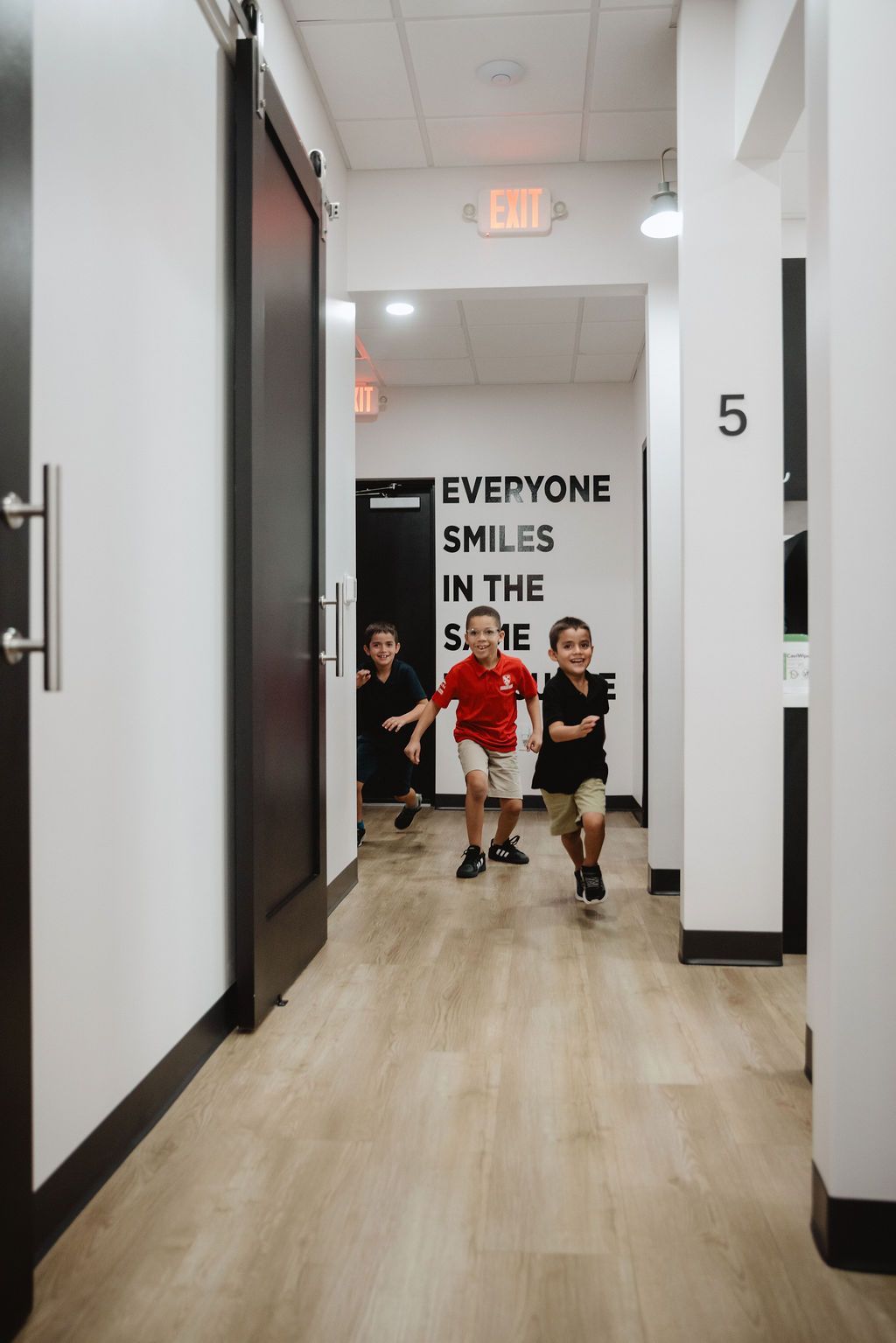 Three children running down a white hallway; black door on the left,