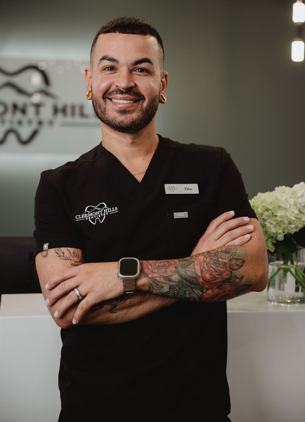 Dentist with arms crossed, smiling. Wearing black scrubs, tattoos, and ear gauges in a dental office.