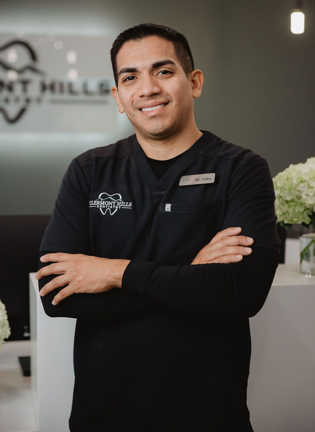 Dentist with arms crossed, smiling, wearing black scrubs, in a dental office.