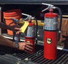 Two red fire extinguishers on a shelf, one being inspected with a tag attached.