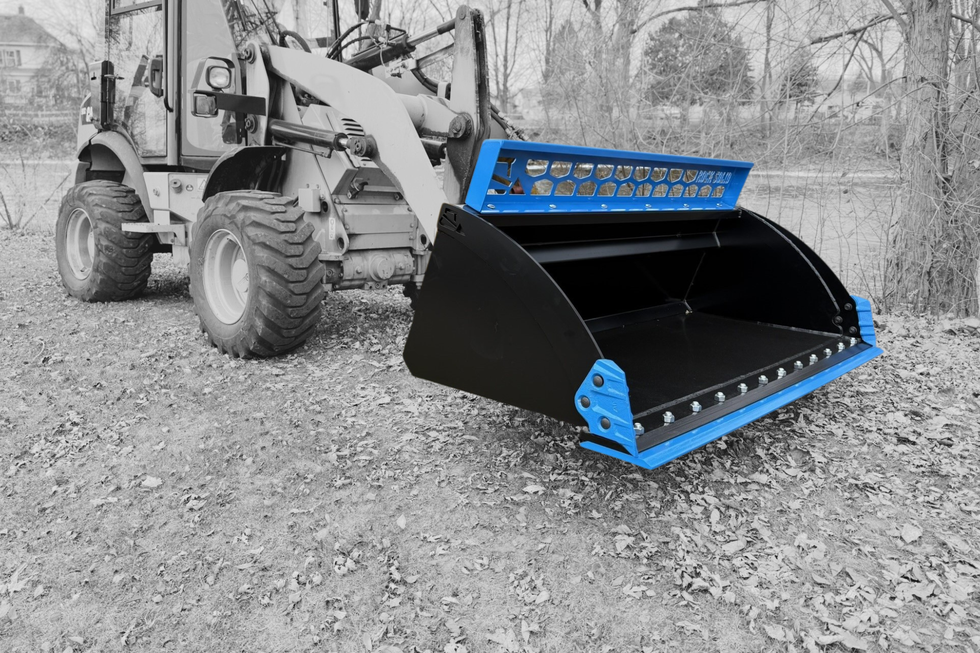Black and blue loader bucket attached to a tractor, outdoors on gravel.
