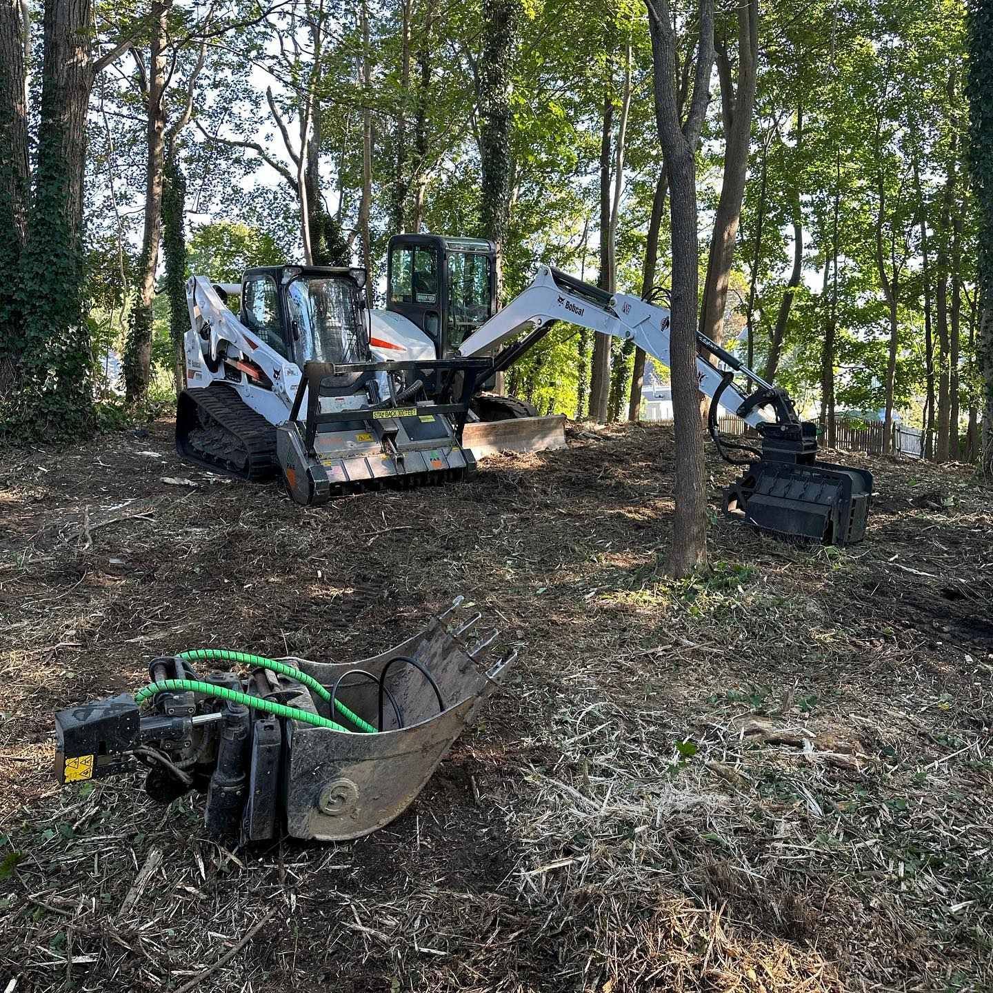 Bobcat excavator in a wooded area with attachments, clearing debris.