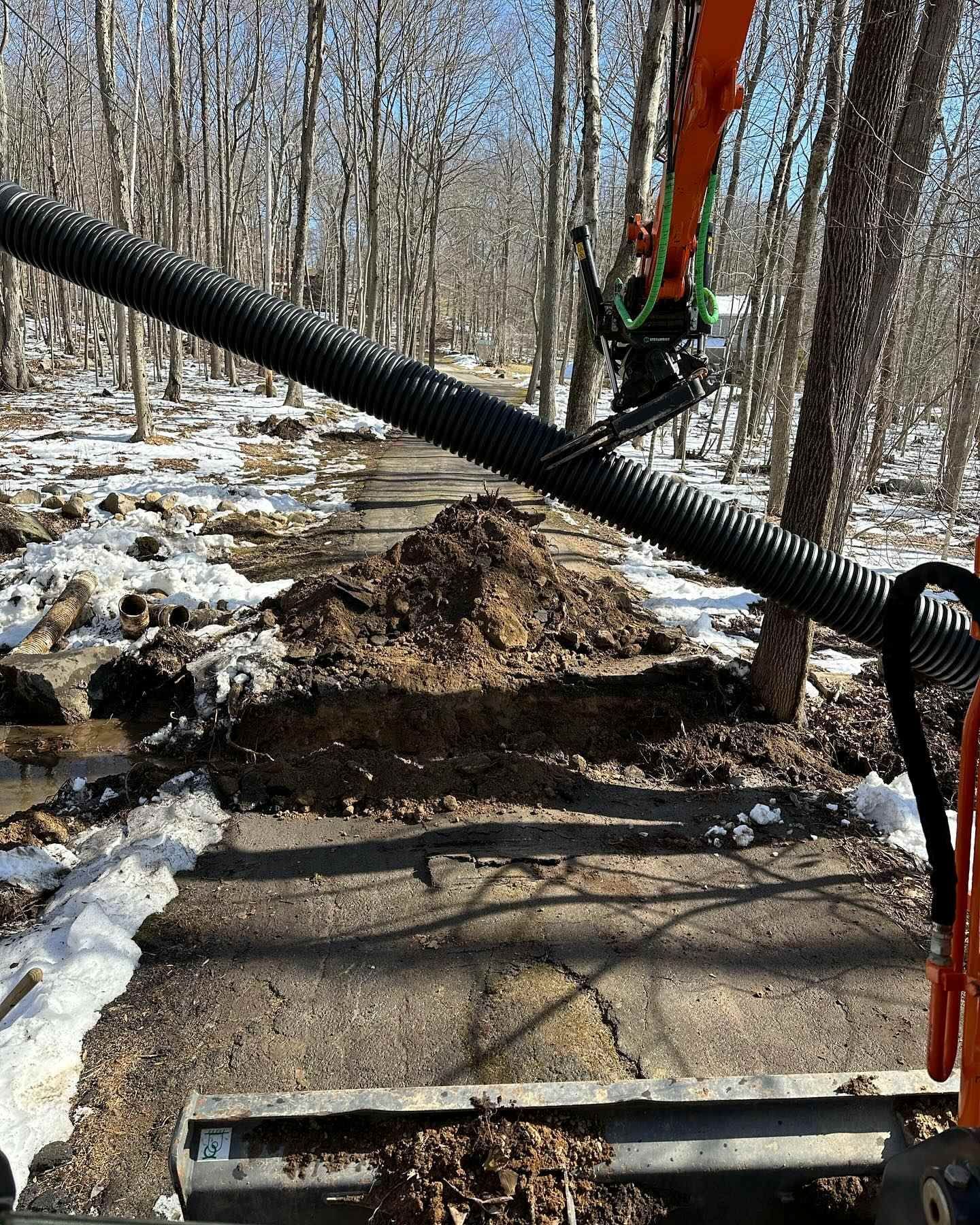 An excavator digging up a path in a wooded area, with black corrugated pipe overhead. Snow and mud are visible.