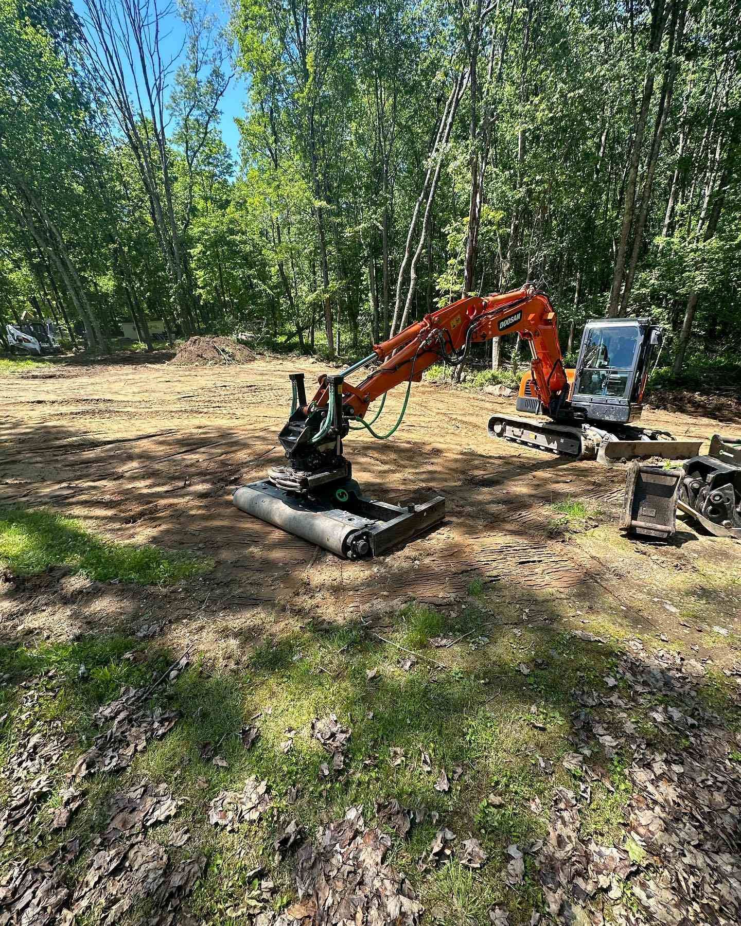 Orange excavator on cleared land, surrounded by trees under a sunny sky.