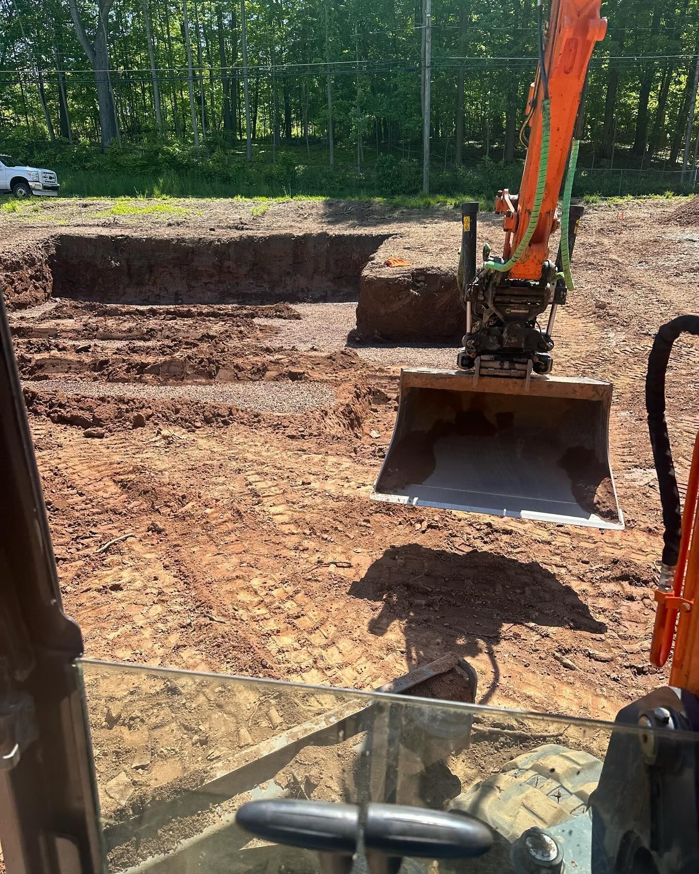 Excavator digging a rectangular foundation pit in a dirt lot. Bright sunlight, orange machinery.