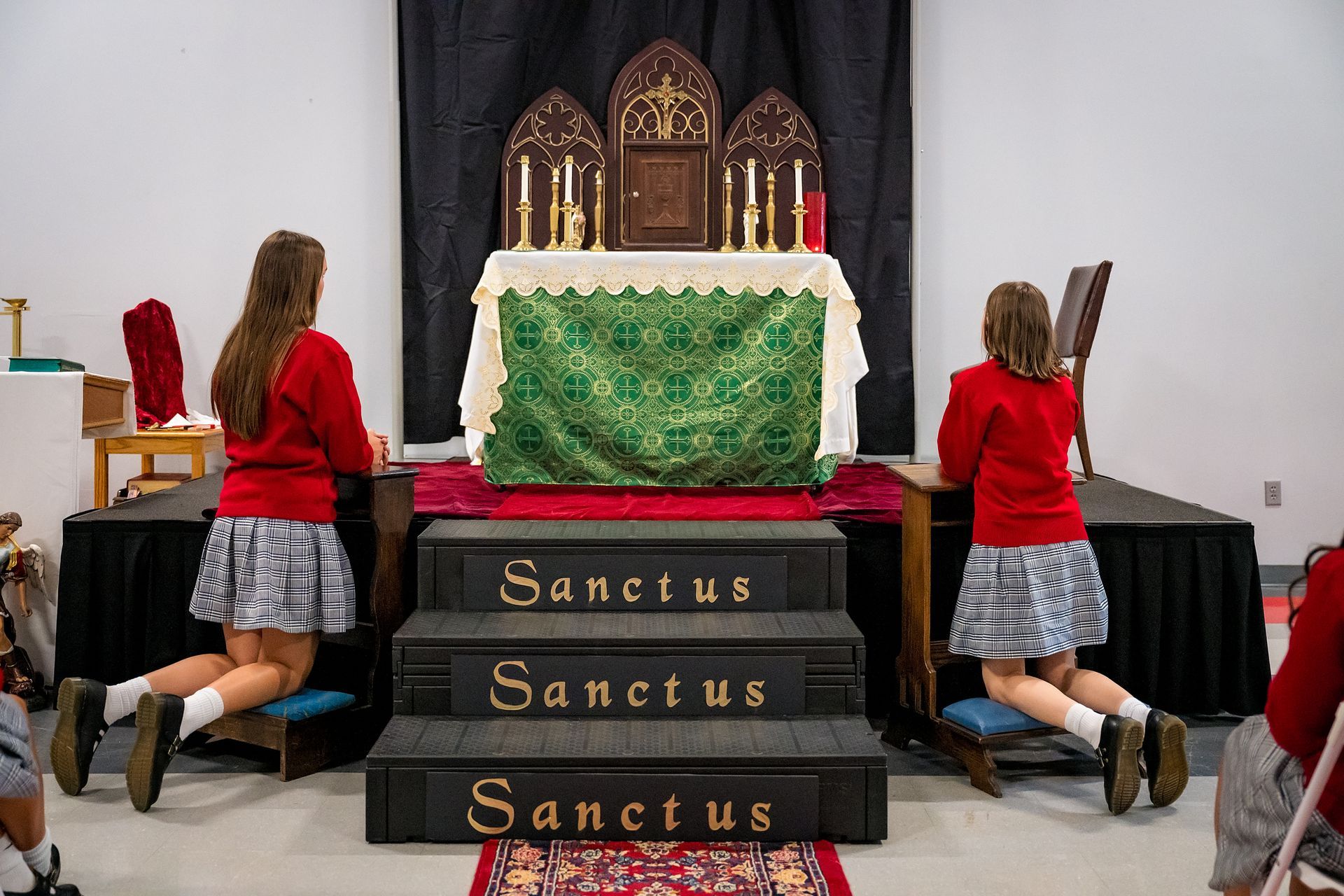 Two kneeling children in red and plaid beside a decorated altar with “Sanctus” on the steps