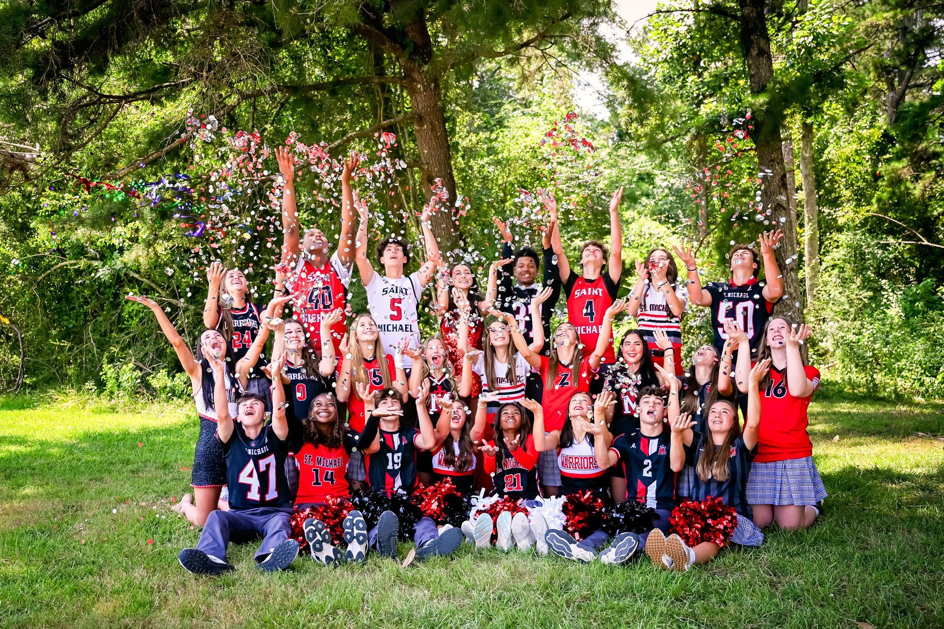 Large group posing outdoors in red, black, and white sports uniforms with arms raised in a leafy park