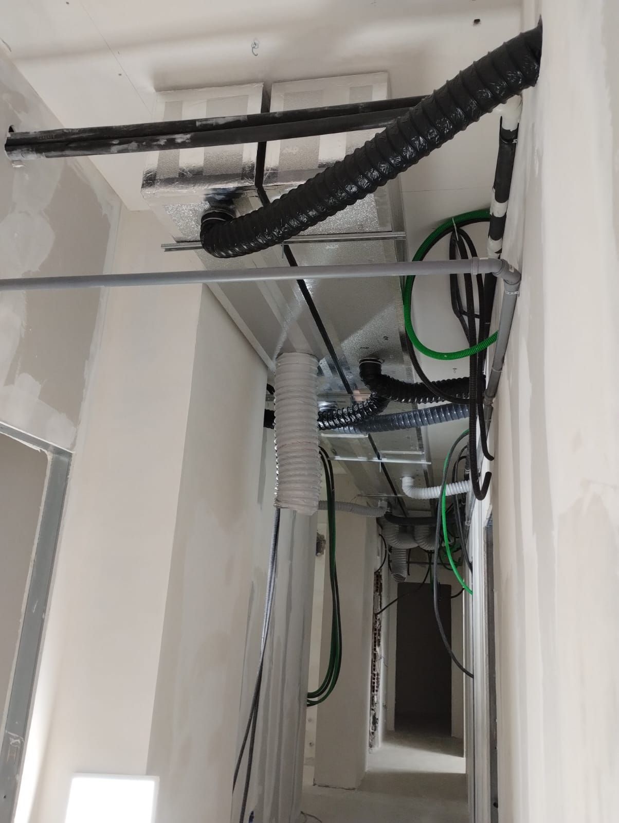 Stretto corridoio di servizio con tubi, cavi e condotti di ventilazione a vista lungo una parete bianca.