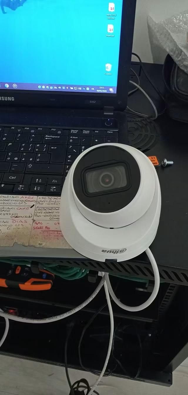 Una telecamera di sicurezza bianca su una scrivania ingombra, accanto a un laptop con schermo blu e cavi.