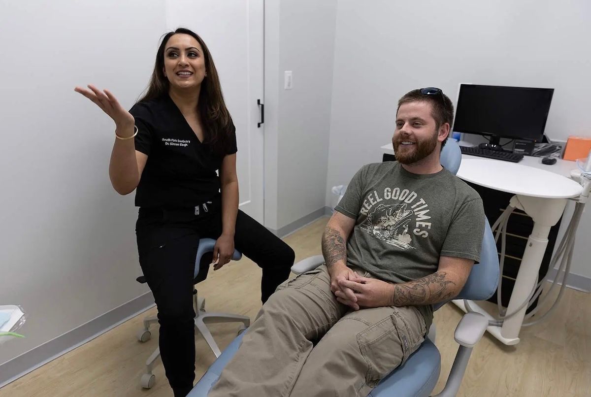 Woman in scrubs pointing at something, sitting in a dentist's office next to a man in a chair.