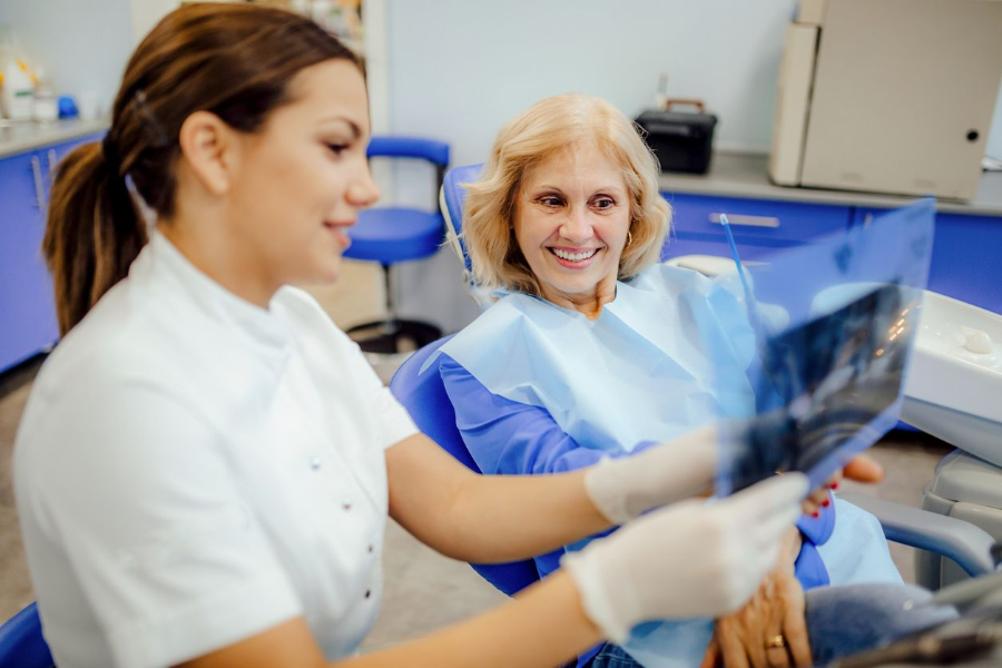 Woman in scrubs pointing at something, sitting in a dentist's office next to a man in a chair.