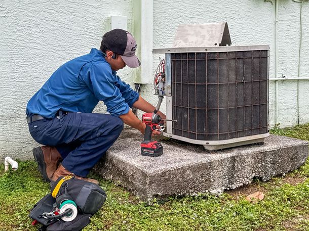 Technician kneels beside an outdoor air conditioner, using a red power drill on the unit.
