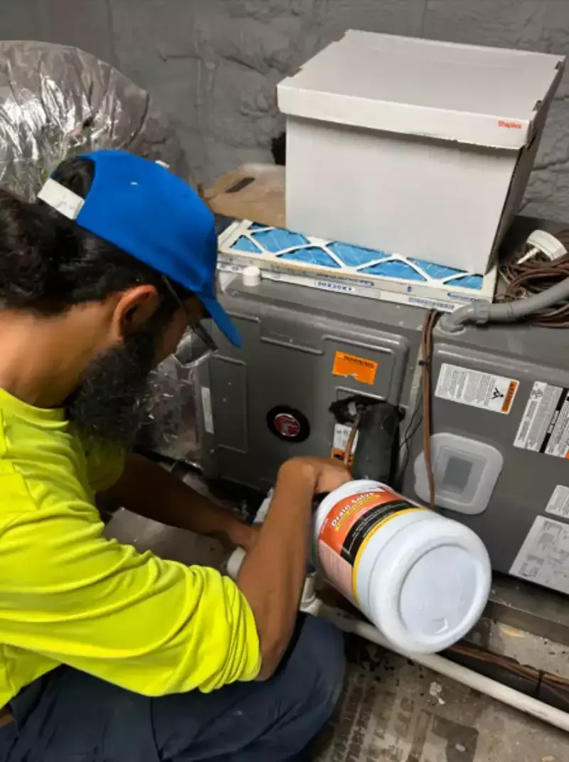 Technician in blue cap repairing HVAC unit with a white cleaner bottle in a utility room