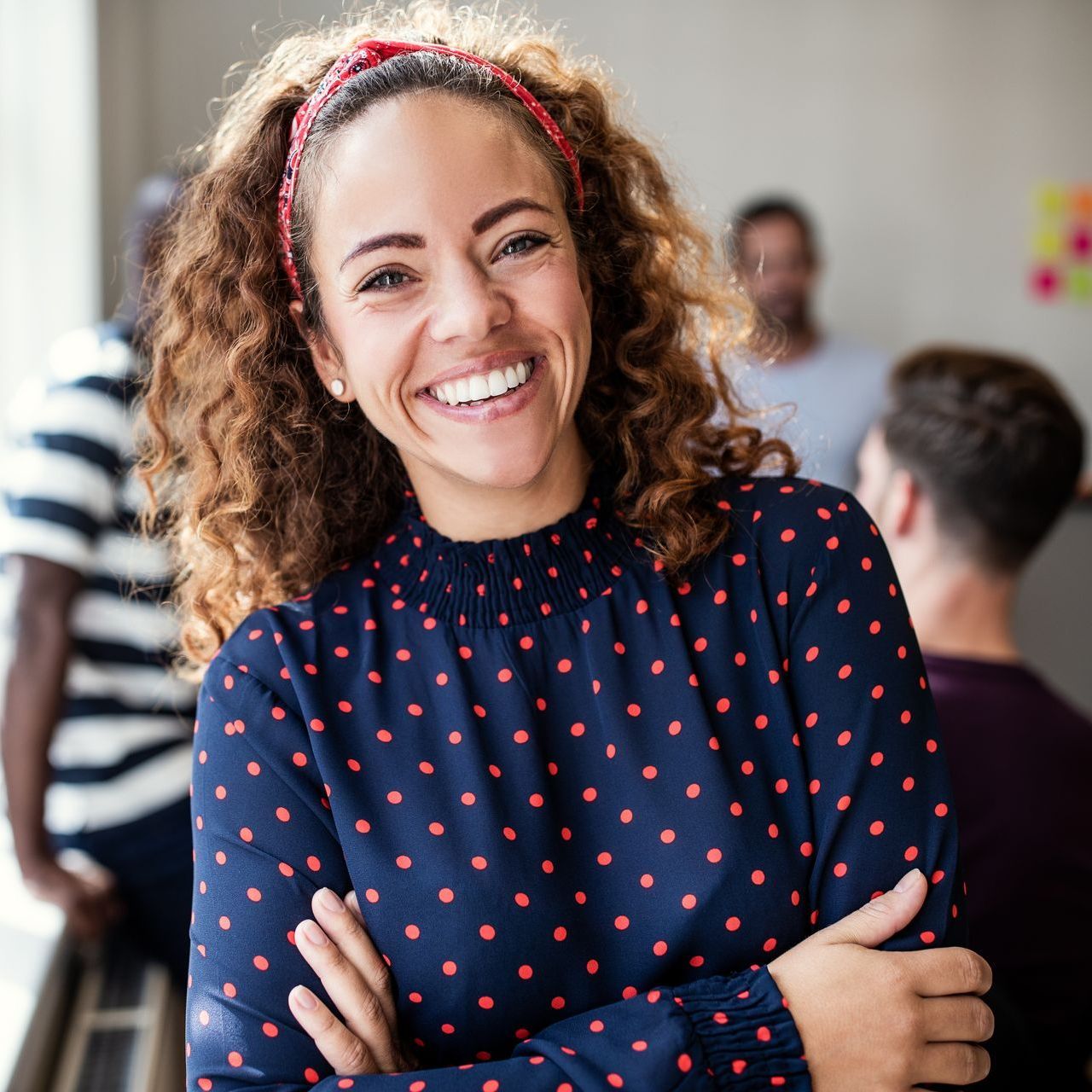 A woman in a blue polka dot shirt is smiling with her arms crossed.