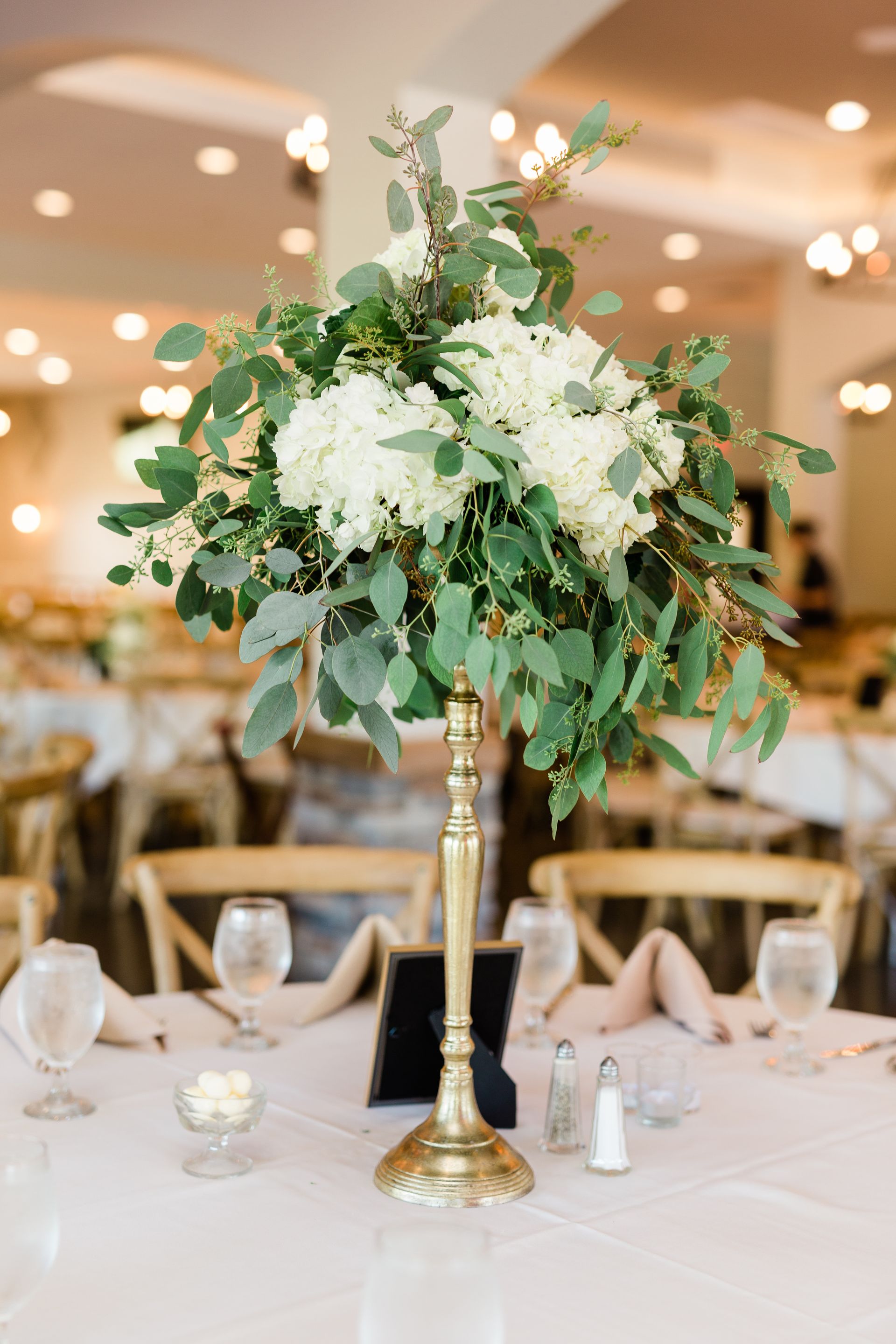A tall gold vase filled with white flowers and greenery is on a table.