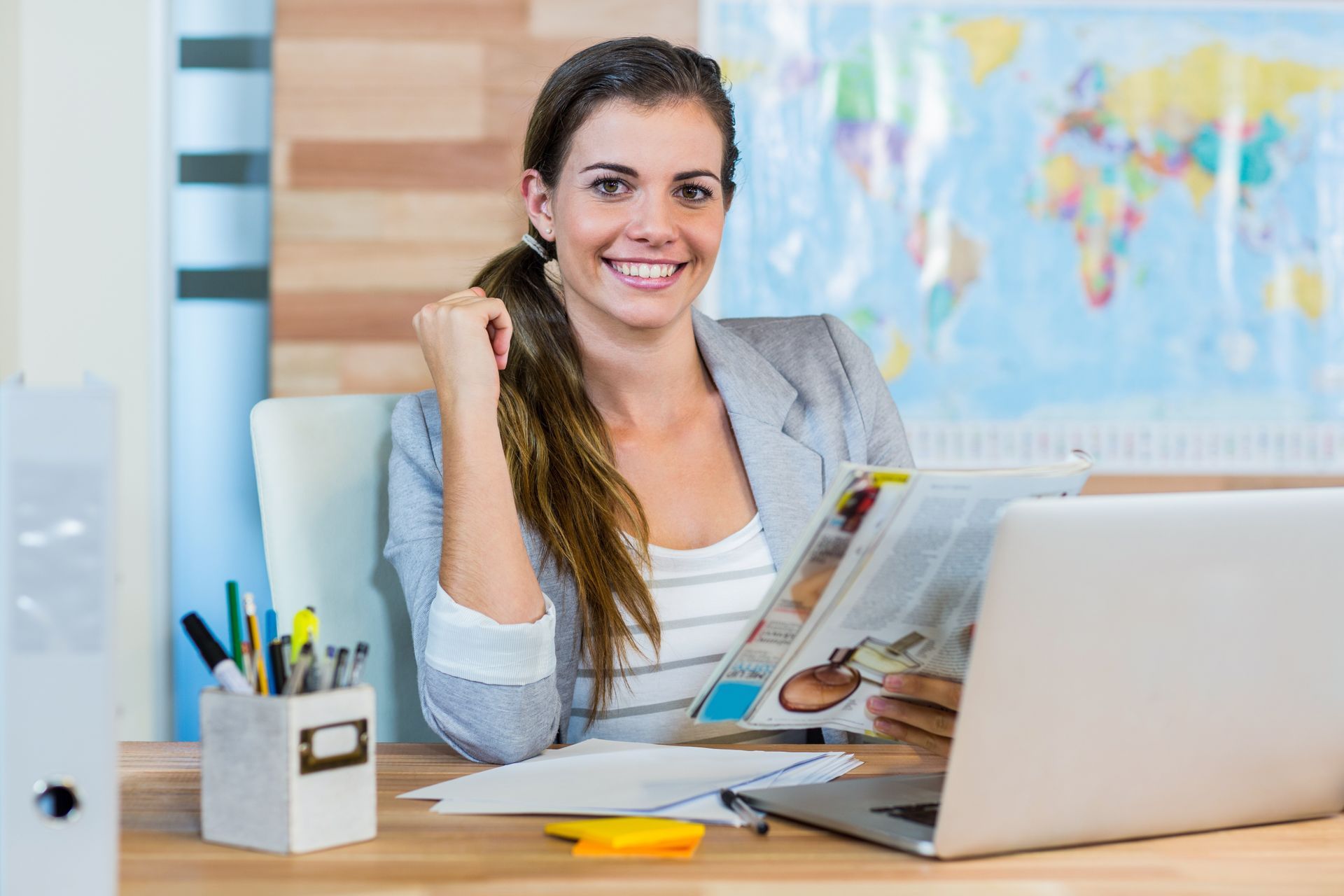 A woman is sitting at a desk with a laptop and a magazine.