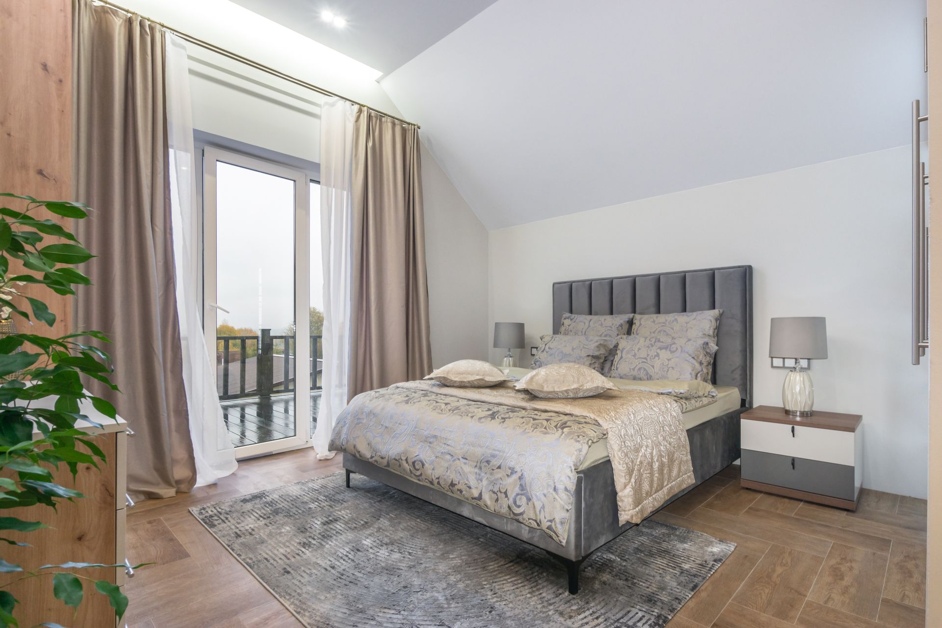 Bright bedroom with gray bed, patterned rug, bedside table, and balcony doors with curtains
