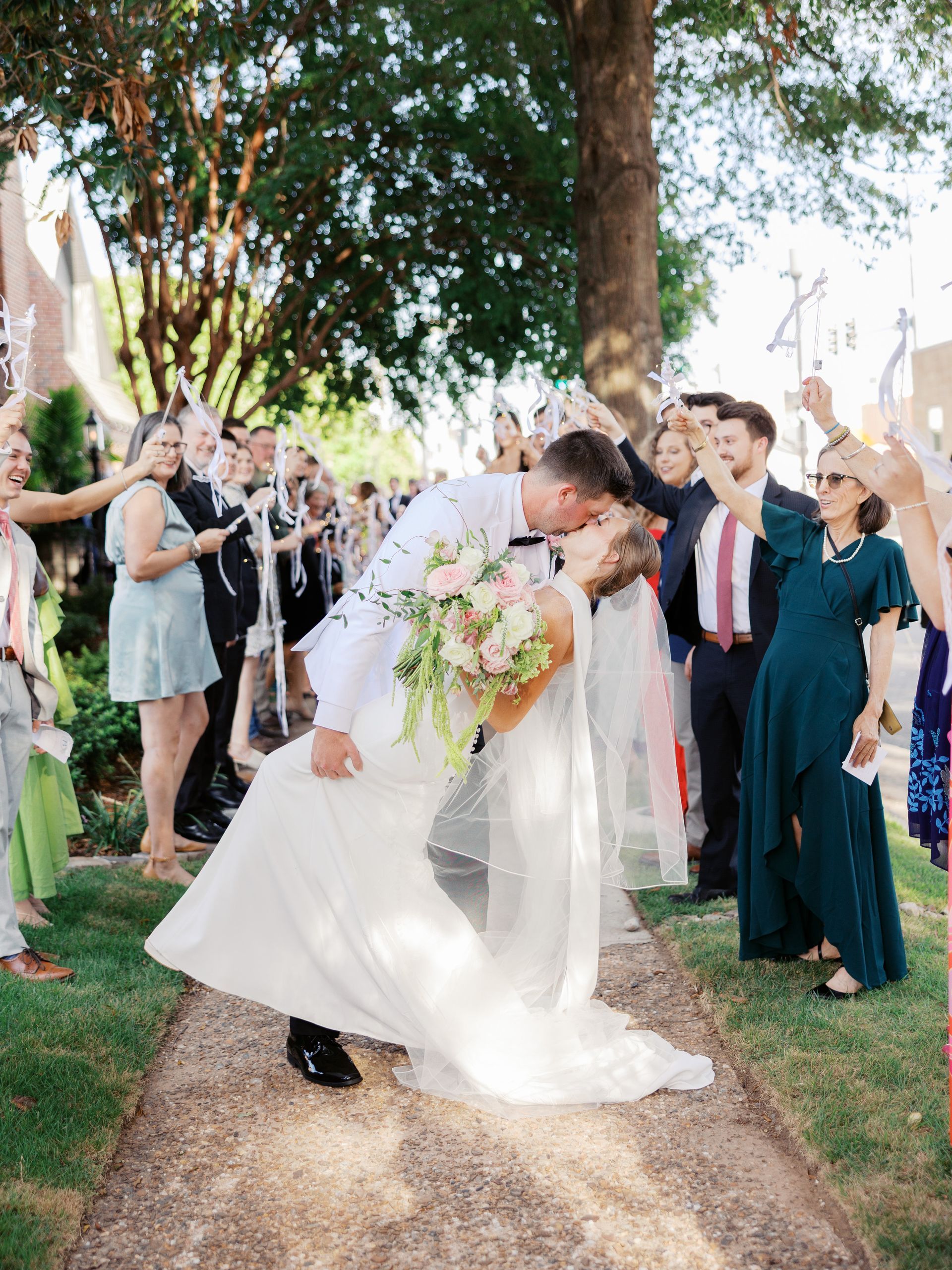 Couple kisses, tilted back, amid confetti shower, guests cheering outside.