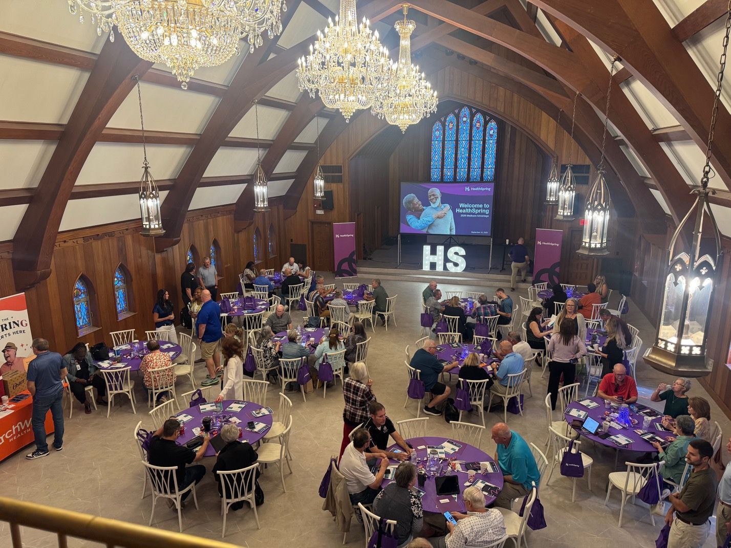 Large event in a high-ceilinged room with tables, people, and a presentation screen. Purple accents.