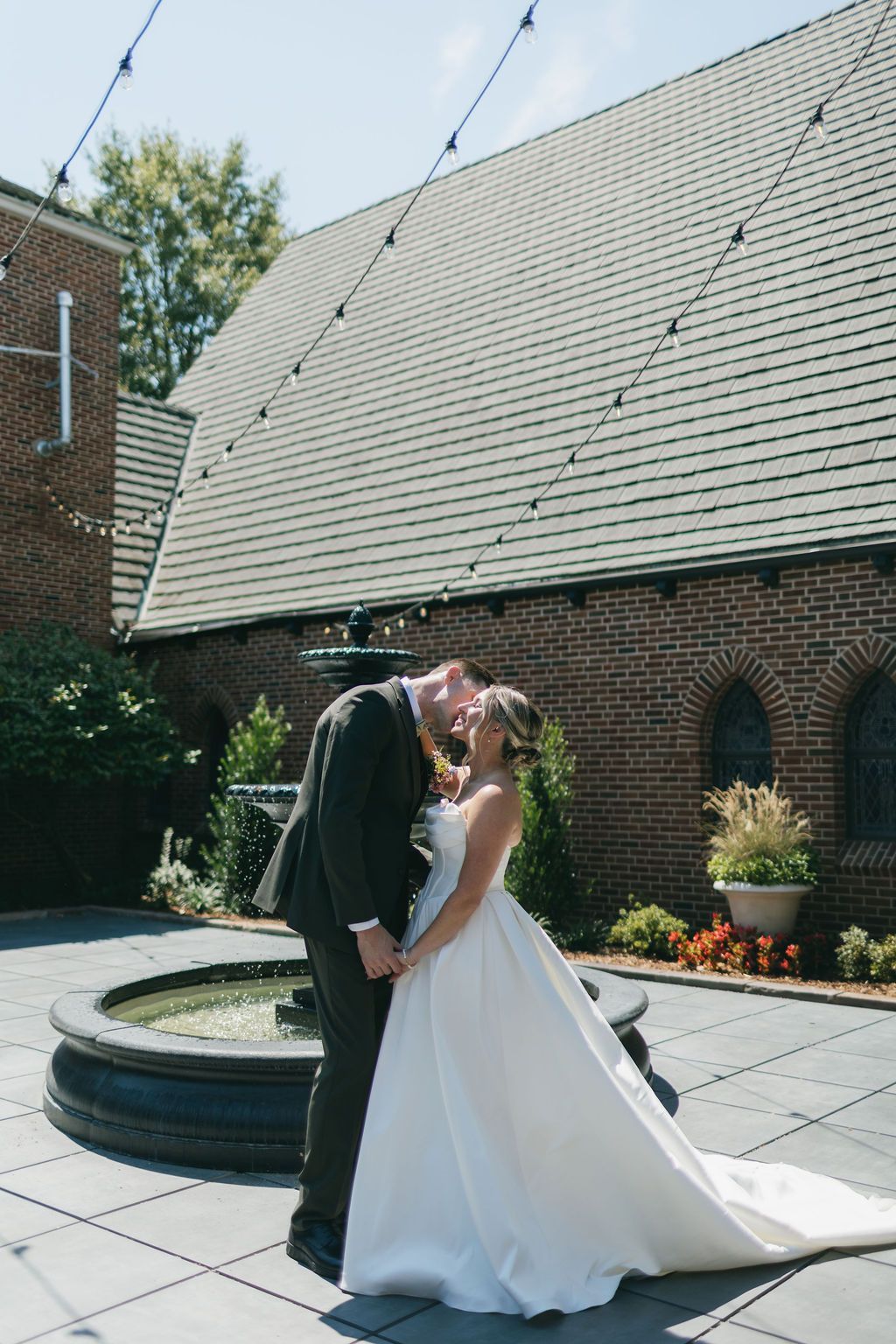Bride in white dress, holding bouquet, poses by a fountain in front of a brick building.