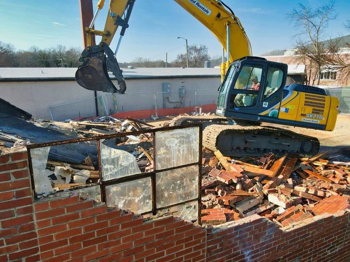 A yellow excavator demolishing a brick building with a large metal claw.