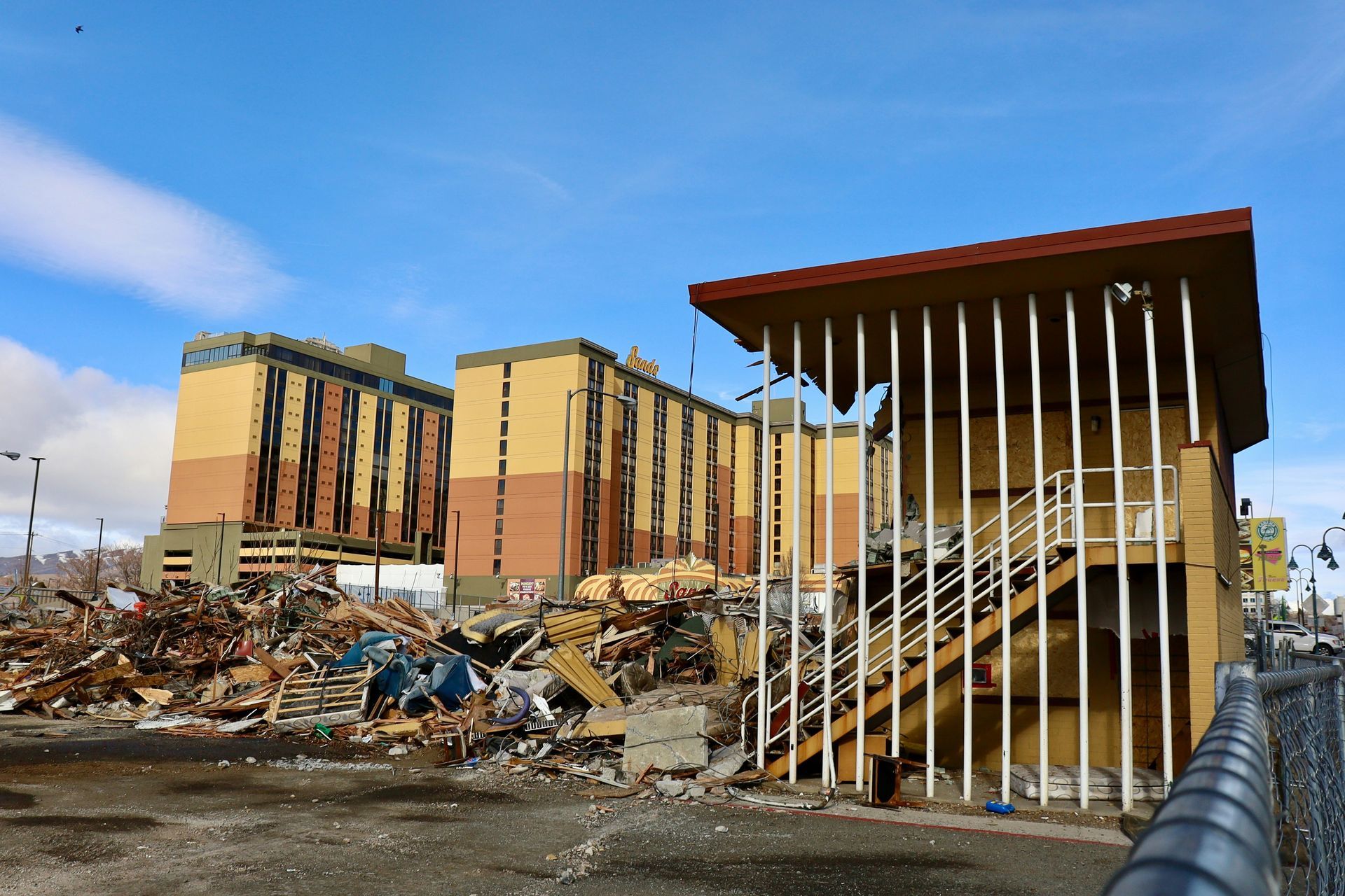 A demolition site showing a pile of rubble in front of a yellow, multi-story building and a small, partially open structure.