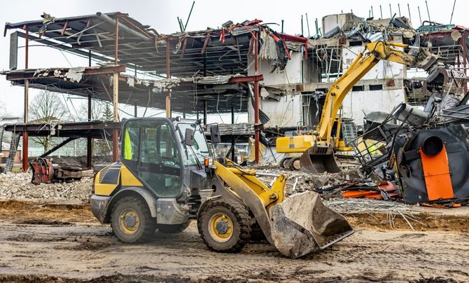 A yellow wheel loader in the foreground and an excavator in the background working at a construction demolition site.