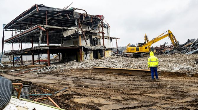 A worker in a yellow high-visibility jacket stands on a muddy site watching an excavator demolish a multi-story building.