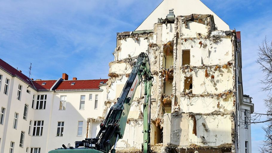 A tall green demolition excavator tears down the side of a multi-story white building against a blue sky.