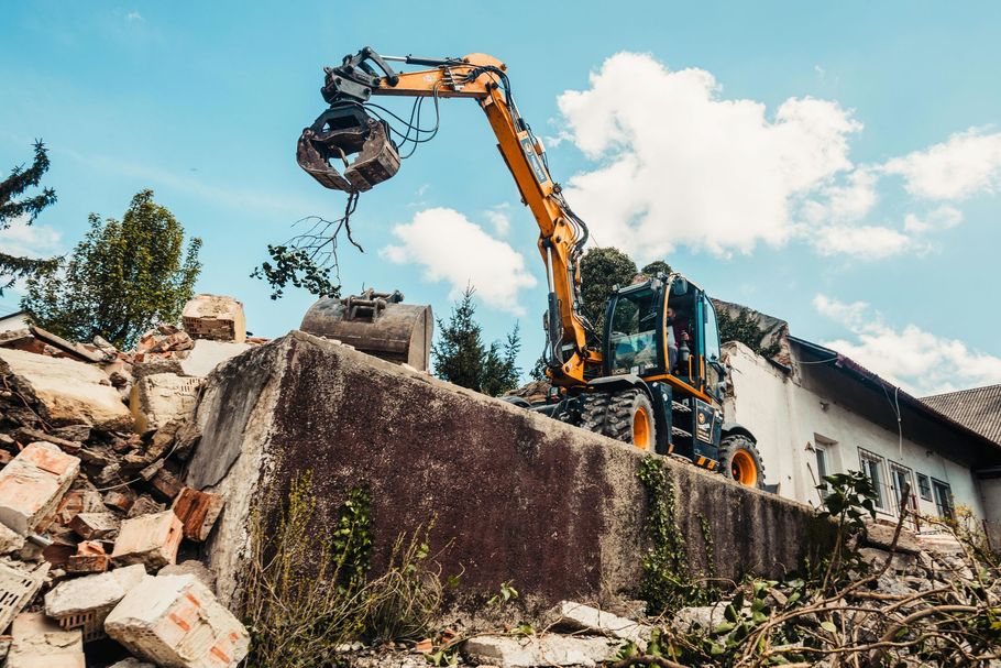 A yellow construction excavator with a claw attachment demolishing a building on a sunny day.
