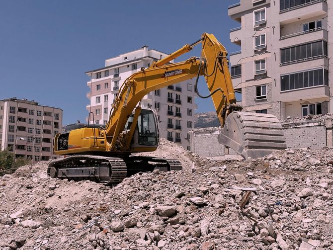 A yellow excavator operates on a pile of concrete rubble in front of multi-story apartment buildings.