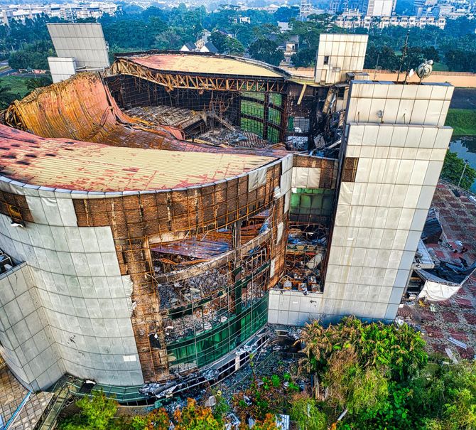 Aerial view of a partially destroyed, charred modern building structure surrounded by green trees and urban development.