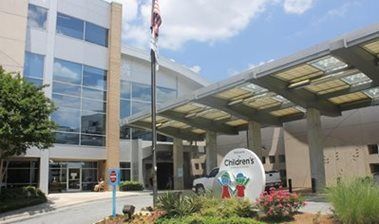 Modern hospital entrance with glass canopy, flagpole, and landscaped flowers outside a building.