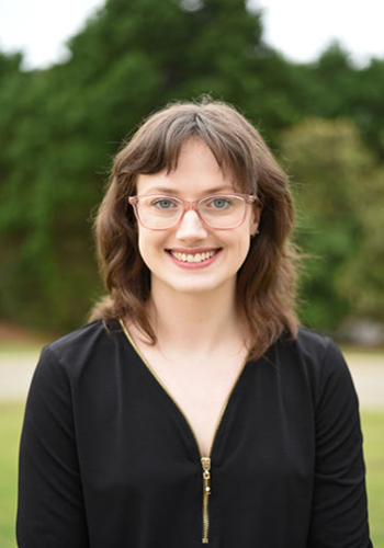 Smiling person in glasses wearing a black top, standing outdoors with blurred trees behind them