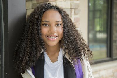 Smiling student with curly hair standing outside a building, wearing a white shirt and purple backpack.